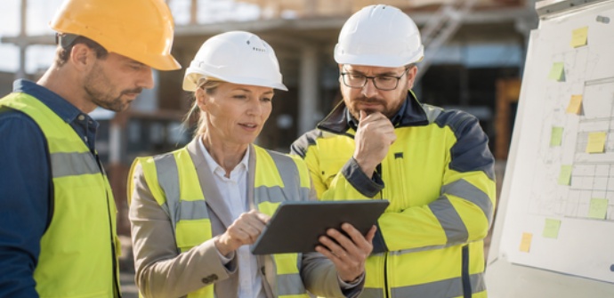 Drei Fachkräfte der Bauwirtschaft besprechen Veränderungen und neue Prozesse auf einer Baustelle. Eine Führungskraft erläutert die nächsten Schritte anhand eines Tablets.
